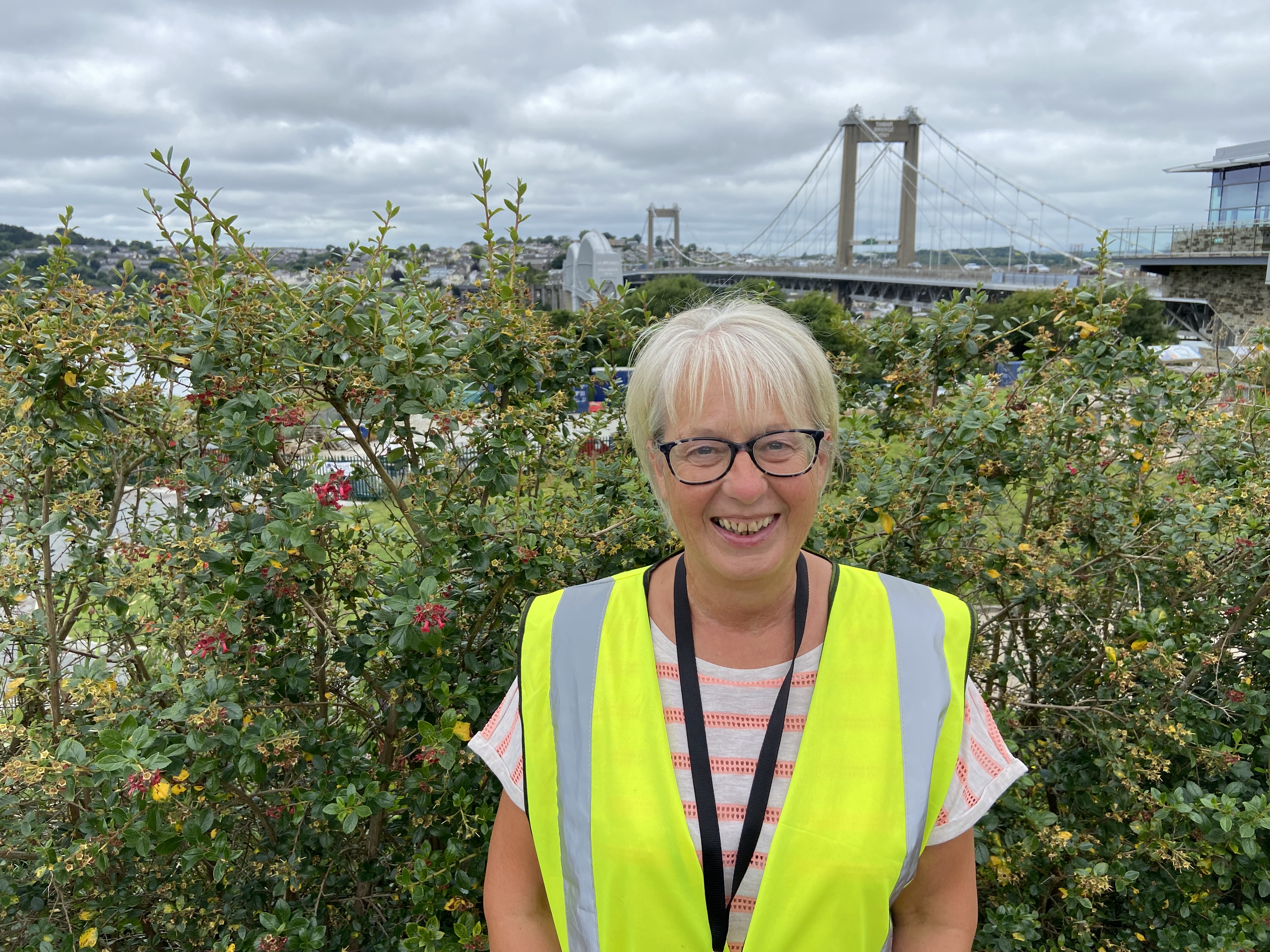 Lady in high vis jacket standing outside in front of greenery and bridges.