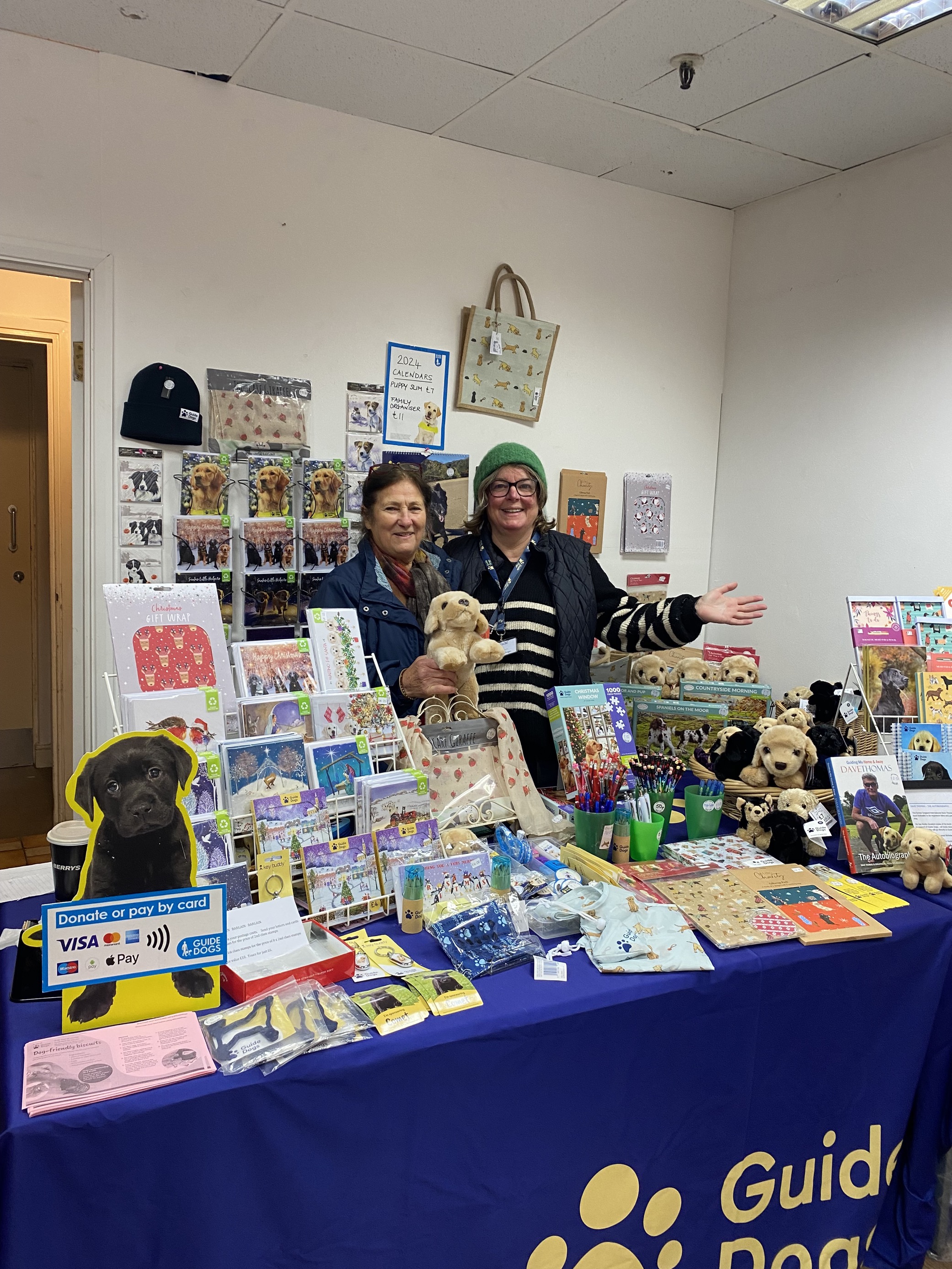 Two volunteers standing behind a desk covered in our Guide Dogs Merchandise. 