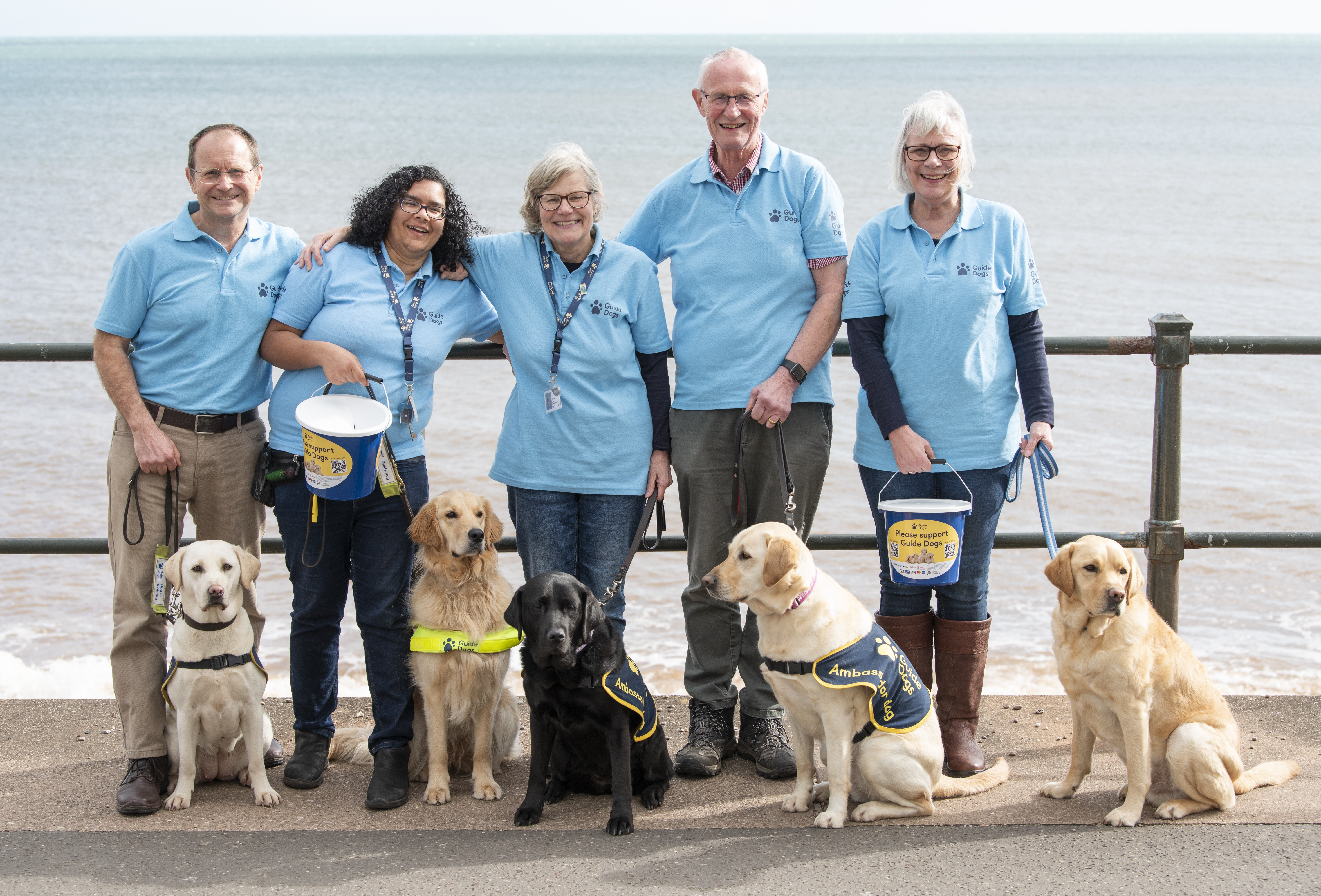 An image of a Fundraising Group with 5 volunteers and several guide dogs. All smiling and looking very happy, holding collection buckets on the beach.