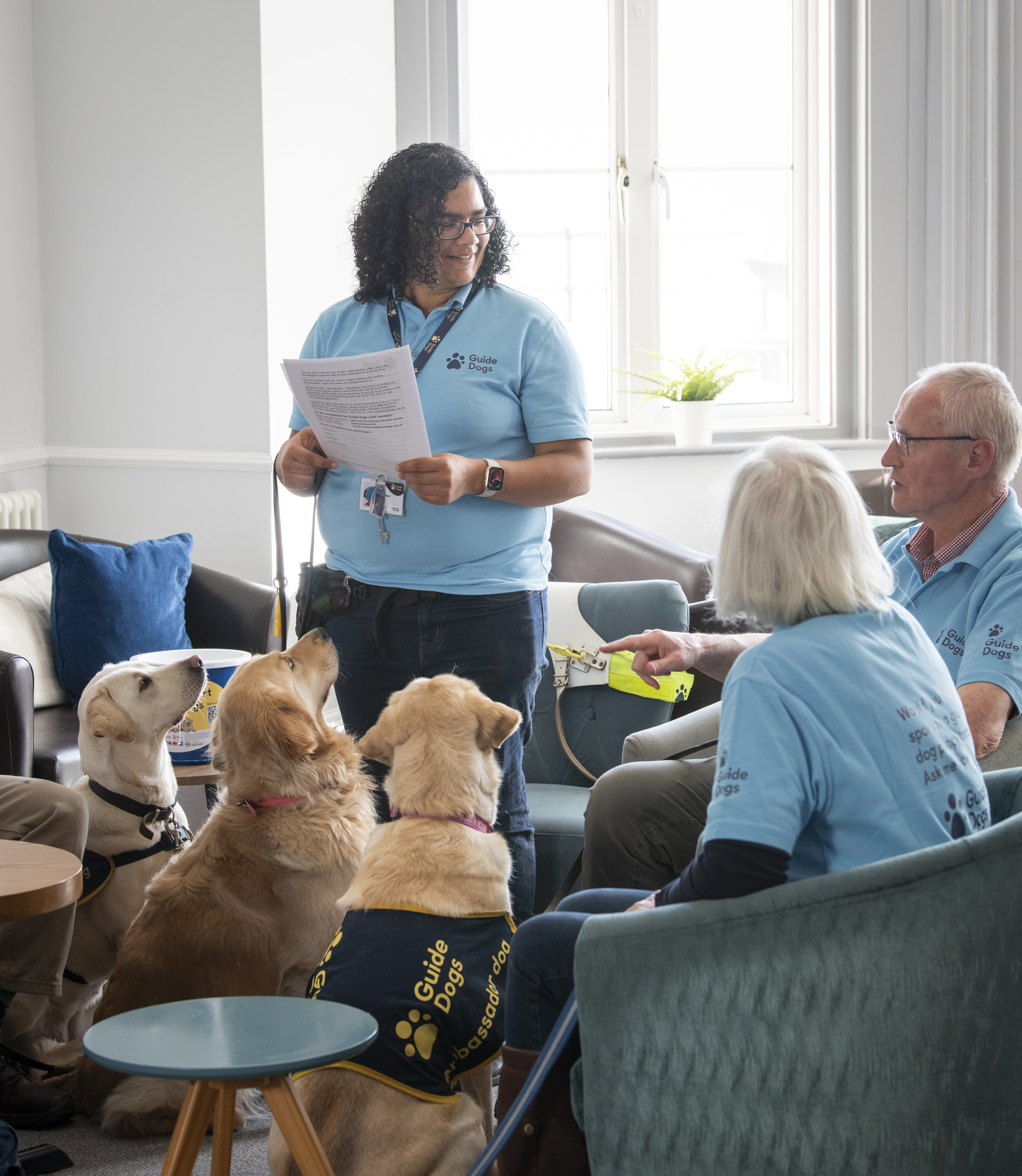 An image of a Fundraising Group Coordinator standing up and talking to their group, with 3 guide dogs looking up at them and listening intently. 
