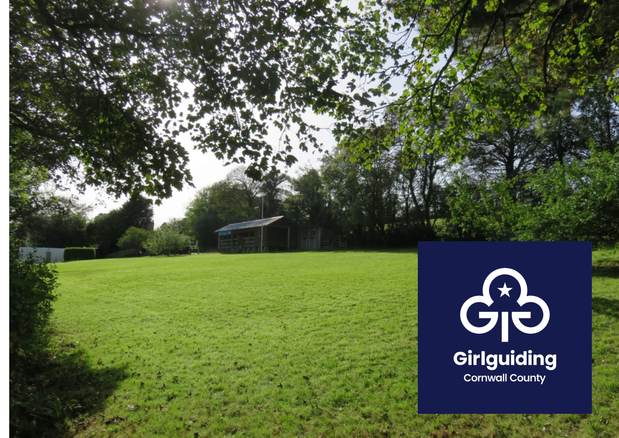 Green field surrounded by trees, with a wooden cabin in the distance. Girlguiding Cornwall logo in bottom right corner.