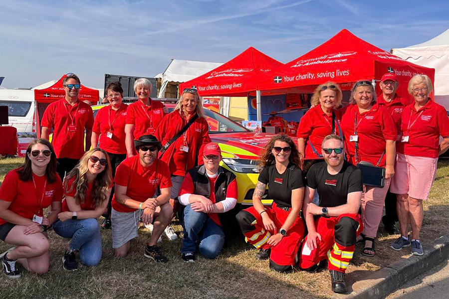 A group fourteen Cornwall Air Ambulance volunteers crouched or standing in front of red event gazebos, and the air ambulance car at a summer event.