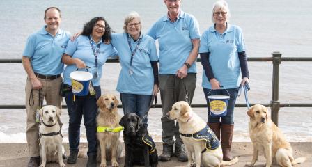 An image of a Fundraising Group with 5 volunteers and several guide dogs. All smiling and looking very happy, holding collection buckets on the beach.