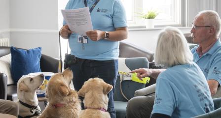An image of a Fundraising Group Coordinator standing up and talking to their group, with 3 guide dogs looking up at them and listening intently. 