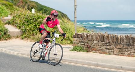 Coast and Clay Cycle Sportive - fundraiser cycling and waving as they pass scenic beach backdrop