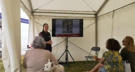 Image of 4 people in an open white gazebo. There is a person standing at the front in a medical detection dogs uniform delivering a talk using a tv screen displaying slides. The other three people are sat on hay bales with their backs to the camera watching along. There is also a white dog who is facing out of the tent.