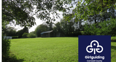 Green field surrounded by trees, with a wooden cabin in the distance. Girlguiding Cornwall logo in bottom right corner.