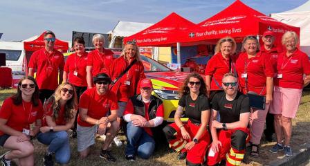 A group fourteen Cornwall Air Ambulance volunteers crouched or standing in front of red event gazebos, and the air ambulance car at a summer event.
